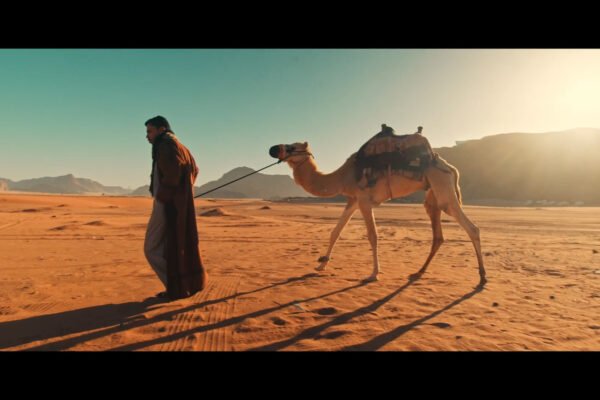 A man walks through a desert with a camel, highlighting the vast landscape and warm sunset tones.