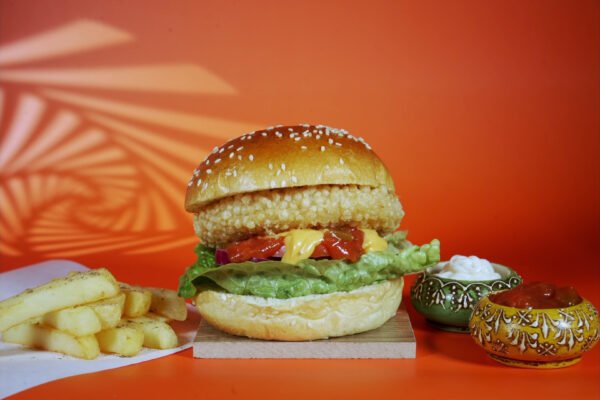 Gourmet burger with lettuce, tomato, and sauce, served with chips and condiments on a vibrant orange background.