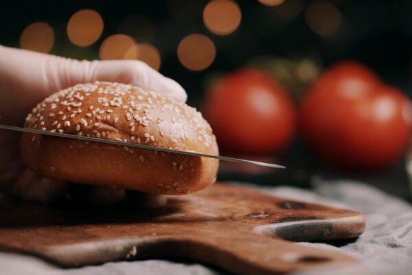 A close-up of a hand slicing a sesame seed burger bun on a wooden board, with tomatoes blurred in the background.