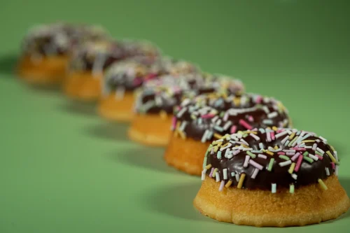 Chocolate doughnuts with colourful sprinkles arranged in a line on a green background, showcasing food photography.