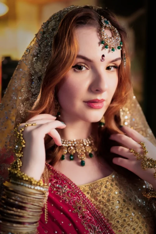 A woman in traditional attire poses elegantly, showcasing intricate jewellery and a vibrant red dupatta against a warm backdrop.