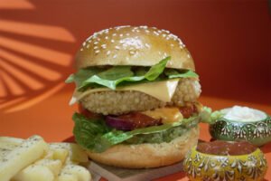 A delicious, layered burger with lettuce, cheese, and tomato, served alongside cheese sticks and sauces on a vibrant orange background.