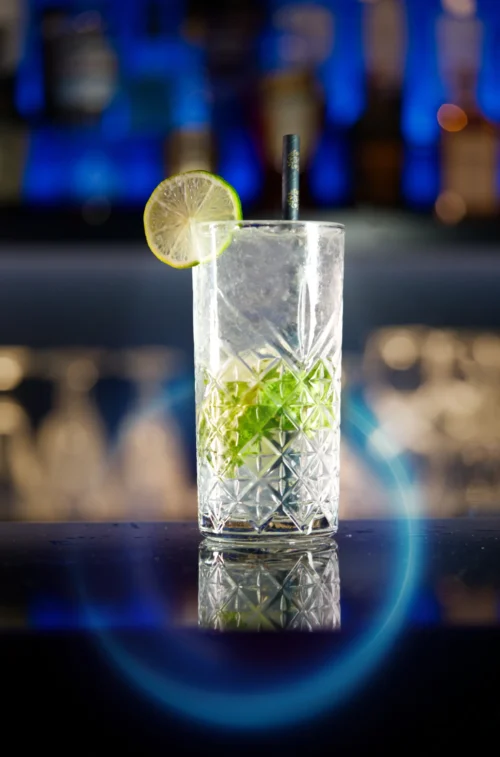 Tall cocktail glass with mint and a lime wedge, reflecting light against a blurred bar backdrop, showcasing enticing food photography.