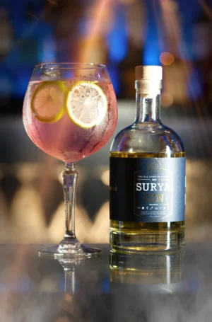A refreshing pink cocktail with lemon slices alongside a bottle of SURYA gin, set against a stylish bar background.